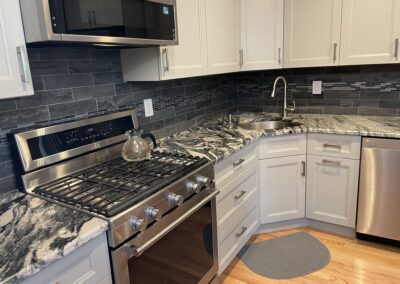 Modern kitchen with a stainless steel gas stove, microwave, and dishwasher. Gray and white granite countertops, white cabinets, and a silver faucet on a wooden floor.