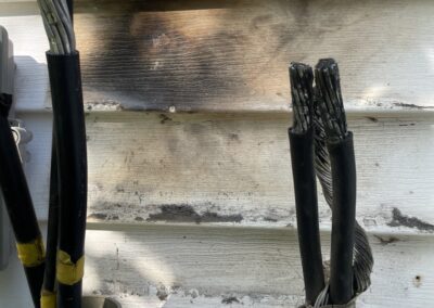Two thick, insulated black cables with exposed wires are mounted on a weathered, white wooden wall. The area shows signs of scorch marks between the cables.