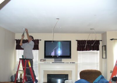Person on a ladder installing ceiling fixtures in a living room with a TV above a fireplace and two windows.